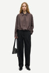 A model stands against a light gray studio background wearing a dark brown textured long-sleeve button-up shirt, black wide-leg trousers, and black shoes, while holding a black shoulder bag.