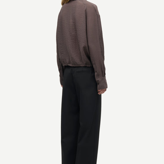 A model shown from the back wearing a dark brown textured long-sleeve shirt and black wide-leg trousers, standing against a light gray background.