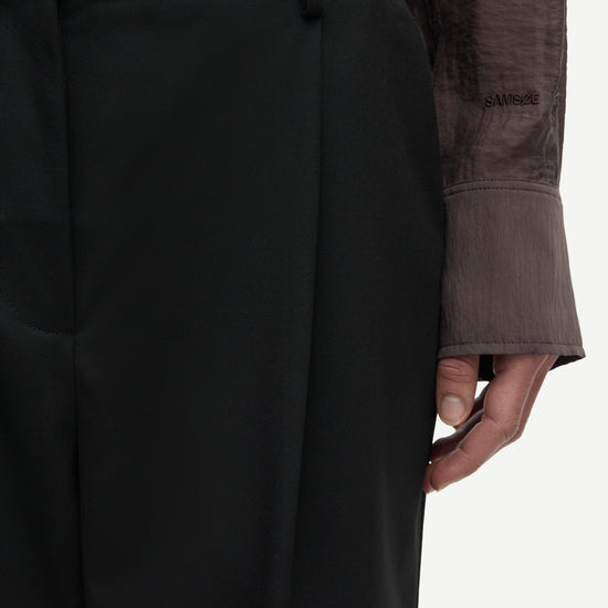 Close-up of a person wearing black high-waisted trousers with a matching belt, paired with a dark sheer long-sleeve top, against a plain light background.