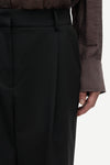 Close-up of a person wearing black high-waisted trousers with a matching belt, paired with a dark sheer long-sleeve top, against a plain light background.