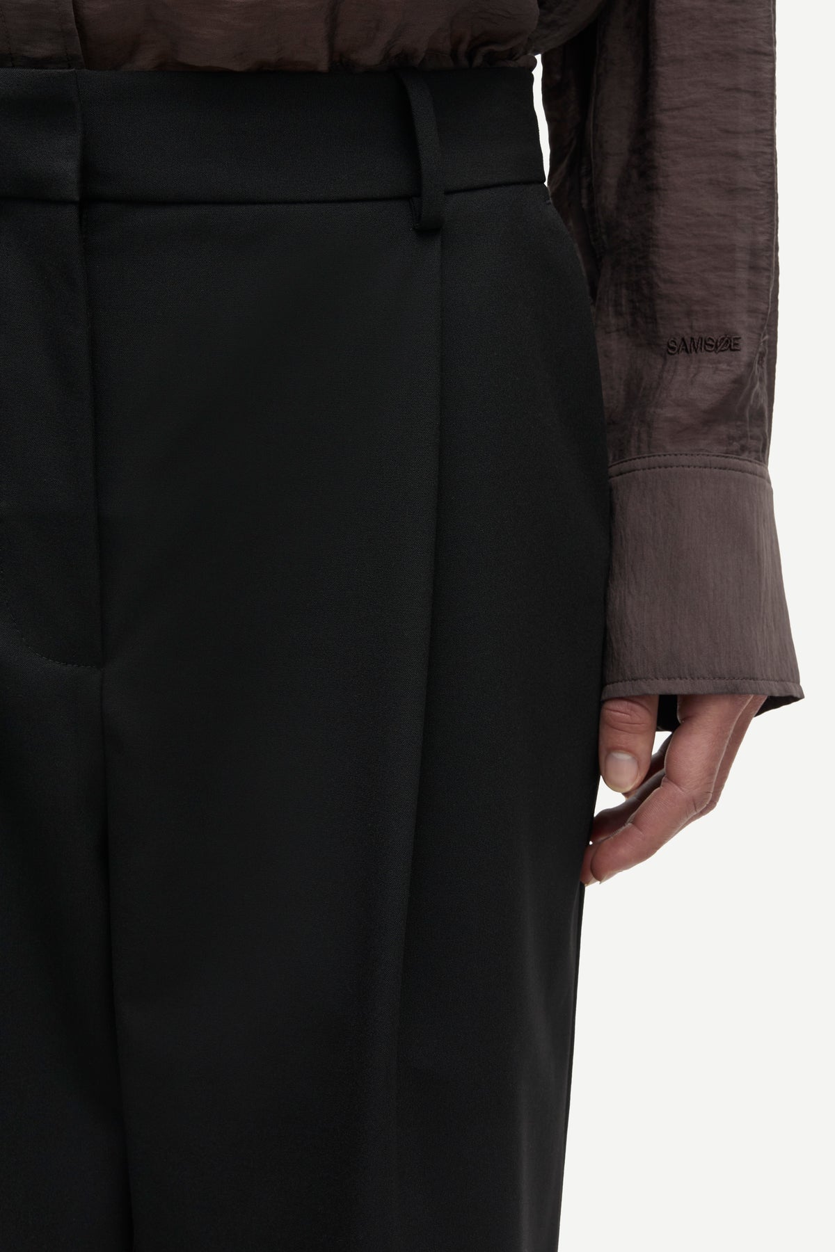 Close-up of a person wearing black high-waisted trousers with a matching belt, paired with a dark sheer long-sleeve top, against a plain light background.