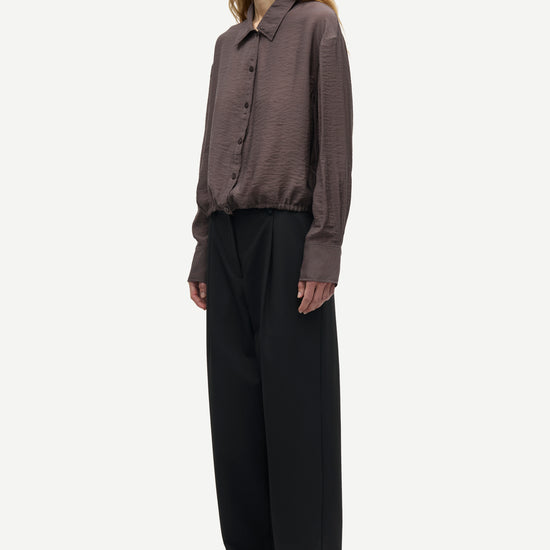 A model shown in profile wearing a dark brown textured long-sleeve button-up shirt and black wide-leg trousers against a light gray background.