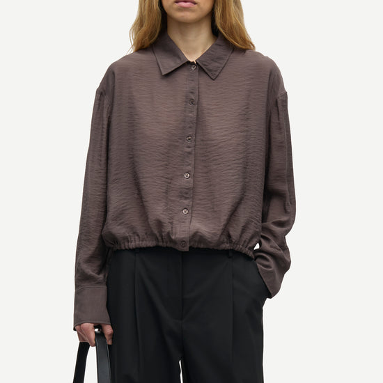 A woman modeling a dark brown crinkled long-sleeve button-up shirt with a pointed collar and gathered hem, paired with black trousers, against a plain light gray background.