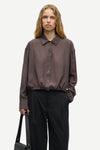 A woman modeling a dark brown crinkled long-sleeve button-up shirt with a pointed collar and gathered hem, paired with black trousers, against a plain light gray background.