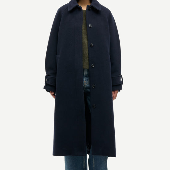 A model wearing a long, dark navy blue wool-blend coat over a green top and blue jeans, standing against a light gray studio background.