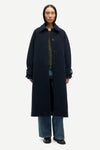 A model wearing a long, dark navy blue wool-blend coat over a green top and blue jeans, standing against a light gray studio background.