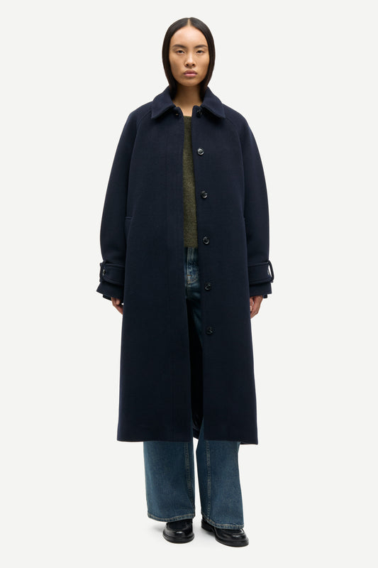 A model wearing a long, dark navy blue wool-blend coat over a green top and blue jeans, standing against a light gray studio background.