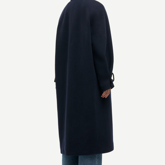 A model shown from the back wearing a long navy blue coat, wide-leg blue jeans, and black shoes against a plain light gray background.
