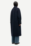 A model shown from the back wearing a long navy blue coat, wide-leg blue jeans, and black shoes against a plain light gray background.