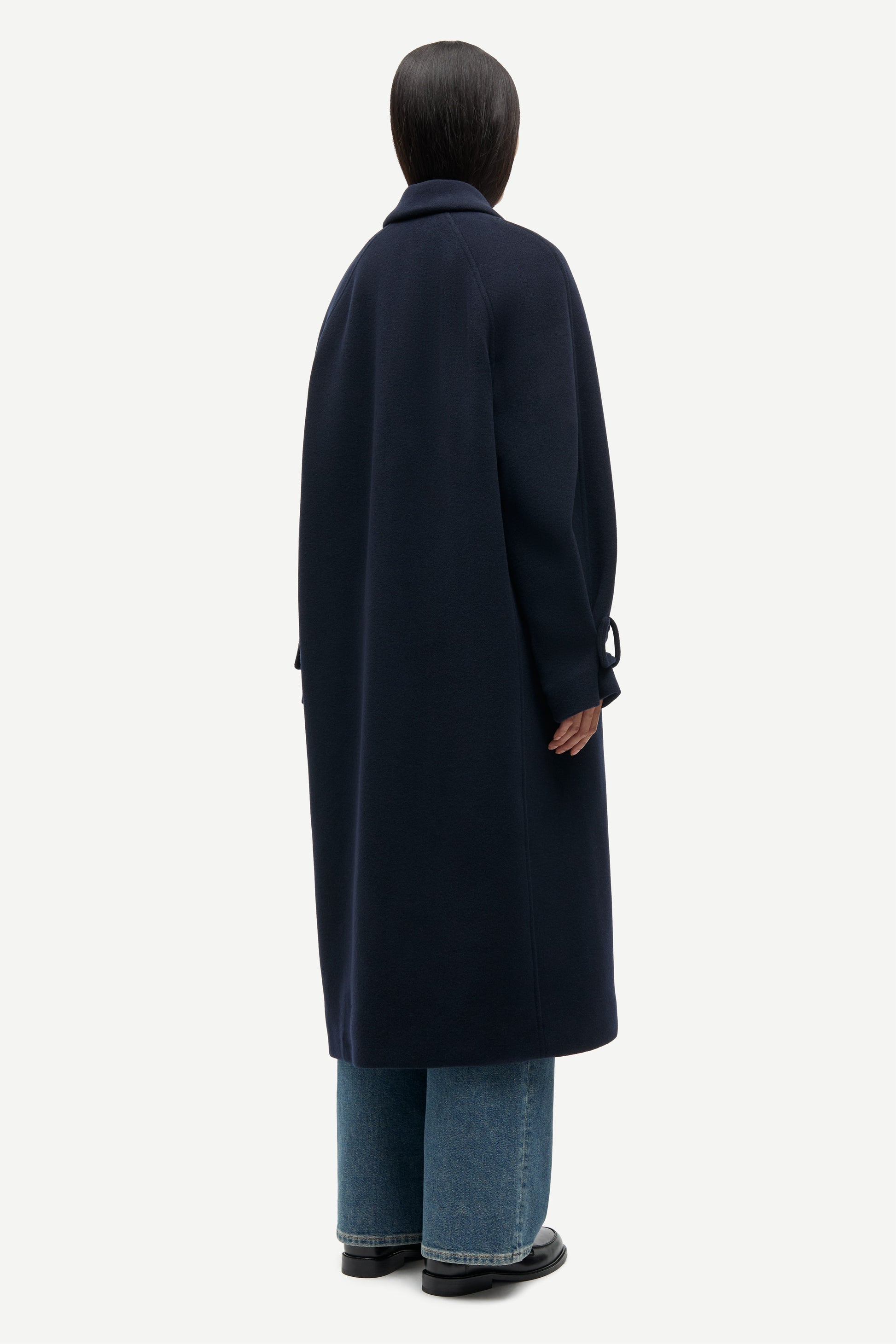A model shown from the back wearing a long navy blue coat, wide-leg blue jeans, and black shoes against a plain light gray background.