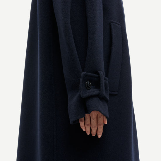 Close-up of a dark navy wool coat with a wide cuff featuring a decorative button detail, worn by a model whose hand is visible.