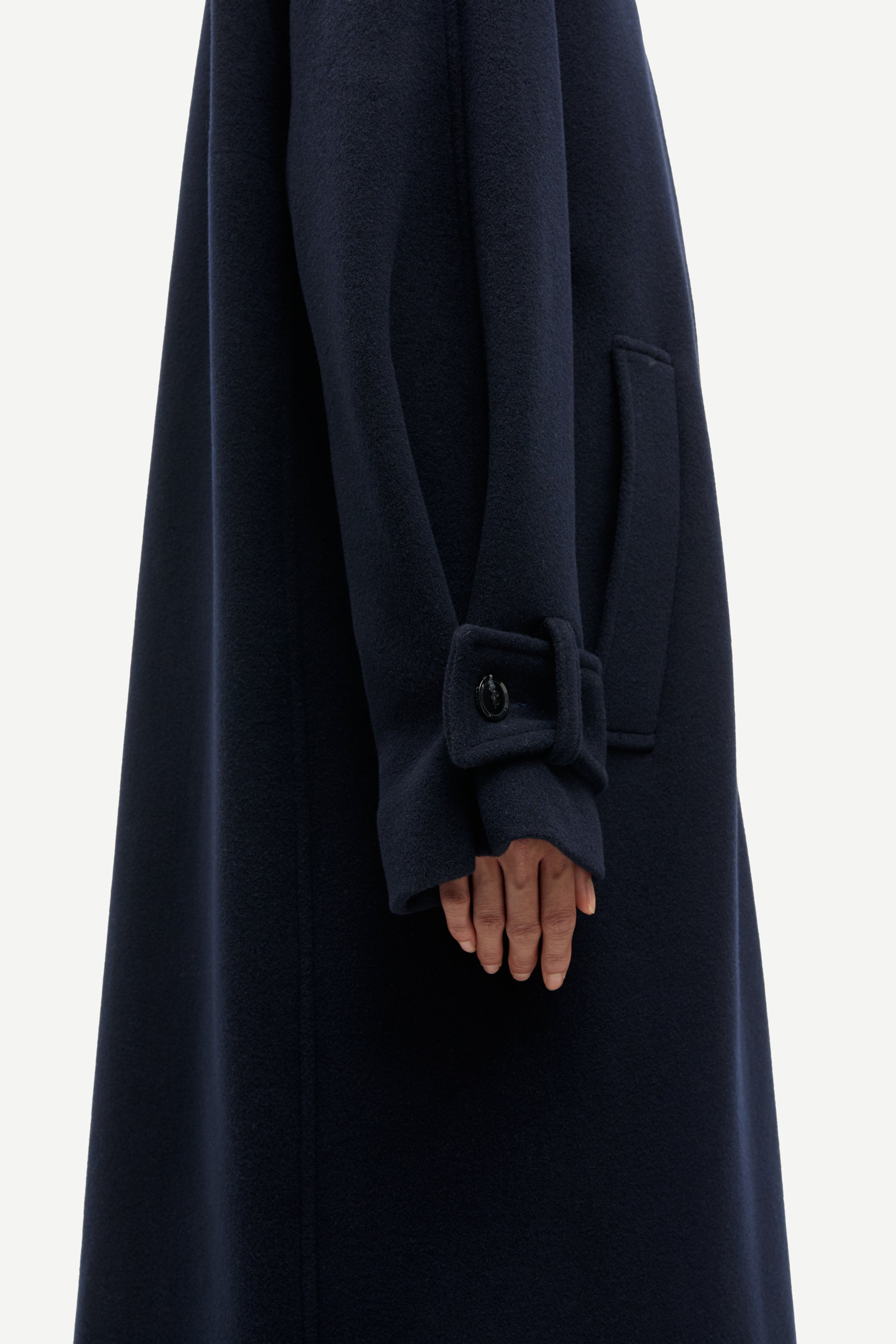 Close-up of a dark navy wool coat with a wide cuff featuring a decorative button detail, worn by a model whose hand is visible.