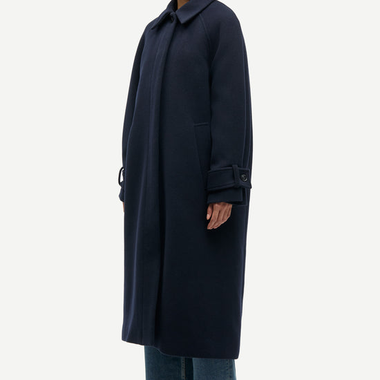 A model wearing a long, dark navy blue wool-blend coat with a collar and buttoned cuffs, styled with wide-leg blue jeans and black shoes, photographed against a solid light gray background.