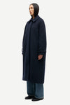 A model wearing a long, dark navy blue wool-blend coat with a collar and buttoned cuffs, styled with wide-leg blue jeans and black shoes, photographed against a solid light gray background.