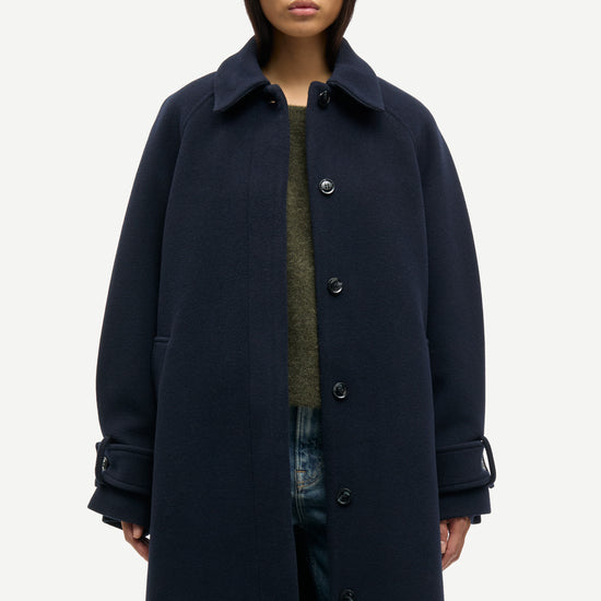 A model wearing a long, dark navy blue wool-blend coat with a wide collar and front buttons, styled over a textured green top and blue jeans, against a plain light gray background.
