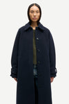 A model wearing a long, dark navy blue wool-blend coat with a wide collar and front buttons, styled over a textured green top and blue jeans, against a plain light gray background.