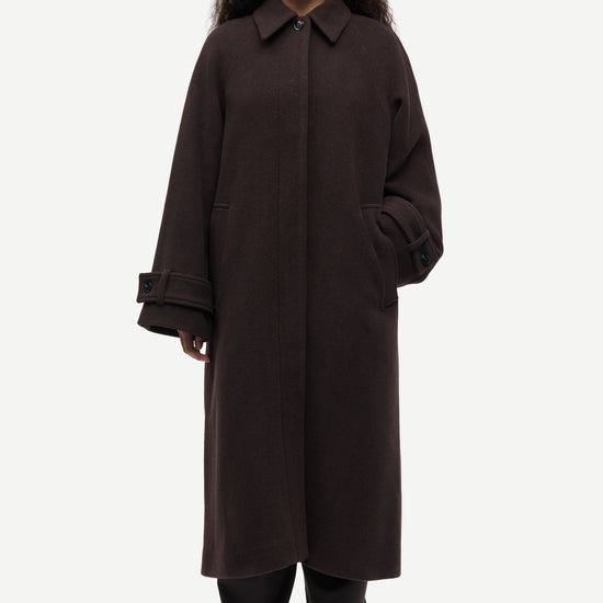 A model wearing a long, dark brown wool-blend coat with a collar and wide sleeves, paired with black trousers and black shoes, against a light gray studio background.