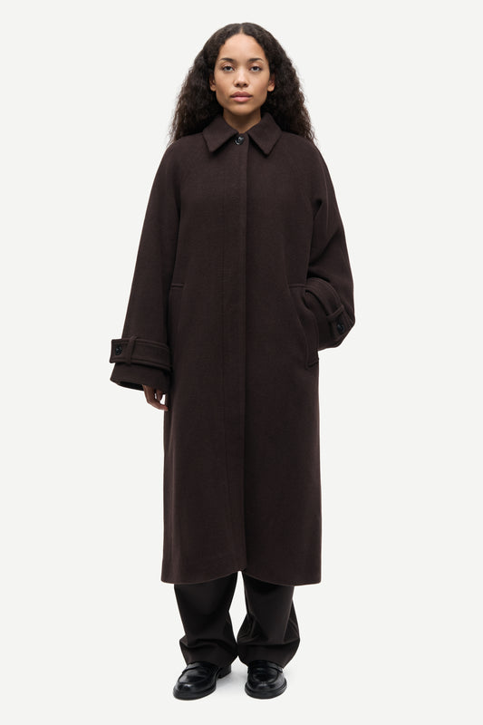 A model wearing a long, dark brown wool-blend coat with a collar and wide sleeves, paired with black trousers and black shoes, against a light gray studio background.