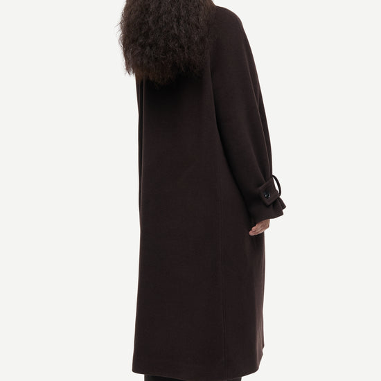 A model shown from the back wearing a long, dark brown wool-blend coat with wide sleeves and tie details at the cuffs, paired with black trousers and shoes, against a light gray studio background.