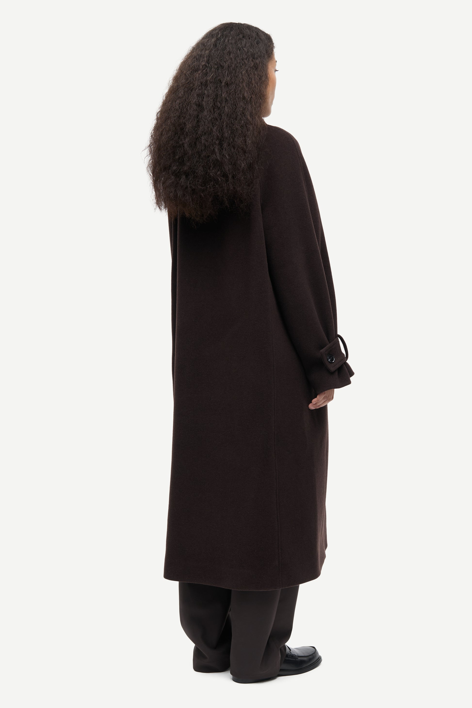 A model shown from the back wearing a long, dark brown wool-blend coat with wide sleeves and tie details at the cuffs, paired with black trousers and shoes, against a light gray studio background.