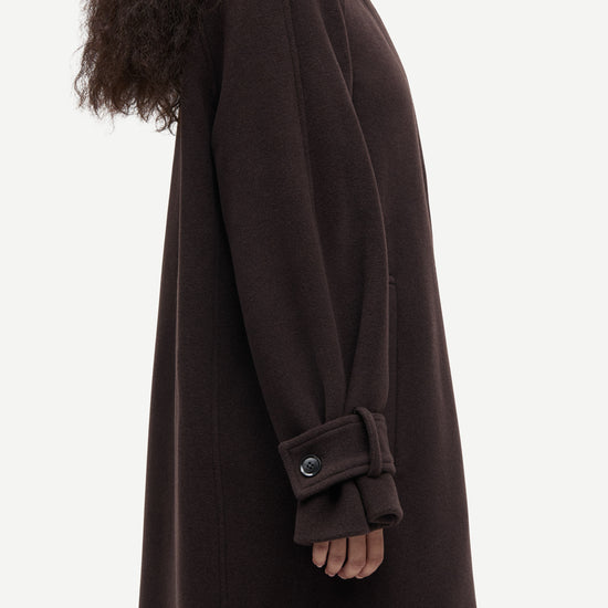A side profile view of a model wearing a long, dark brown wool coat with a collar and a buttoned cuff detail, set against a plain light gray background.