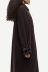A side profile view of a model wearing a long, dark brown wool coat with a collar and a buttoned cuff detail, set against a plain light gray background.