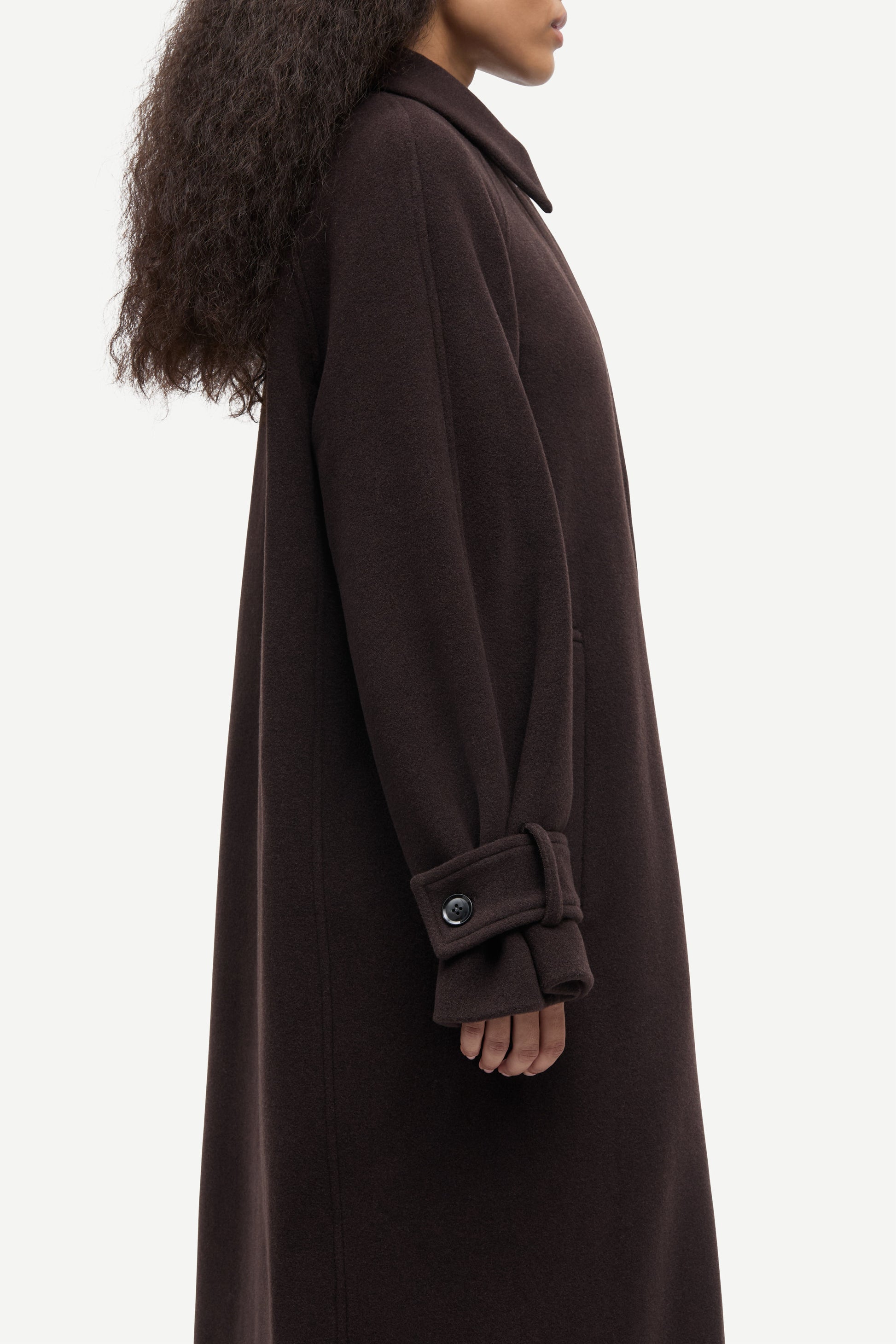 A side profile view of a model wearing a long, dark brown wool coat with a collar and a buttoned cuff detail, set against a plain light gray background.