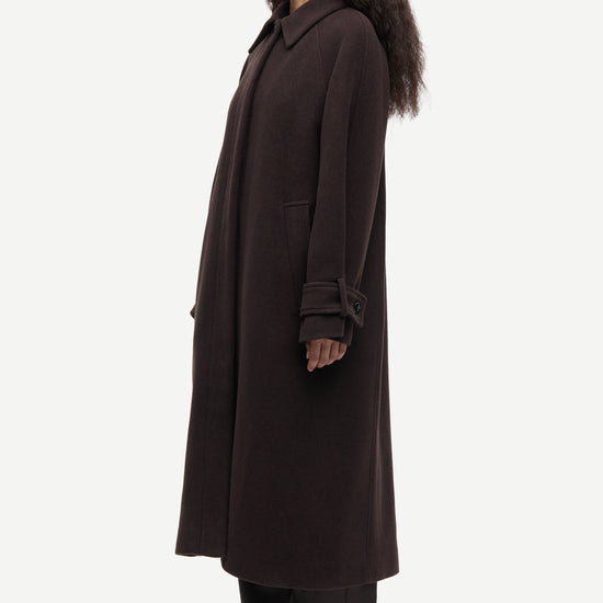 A woman modeling a long, dark brown wool-blend coat with matching dark trousers and black shoes, shown in profile against a light gray studio background.