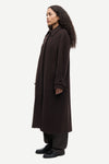 A woman modeling a long, dark brown wool-blend coat with matching dark trousers and black shoes, shown in profile against a light gray studio background.