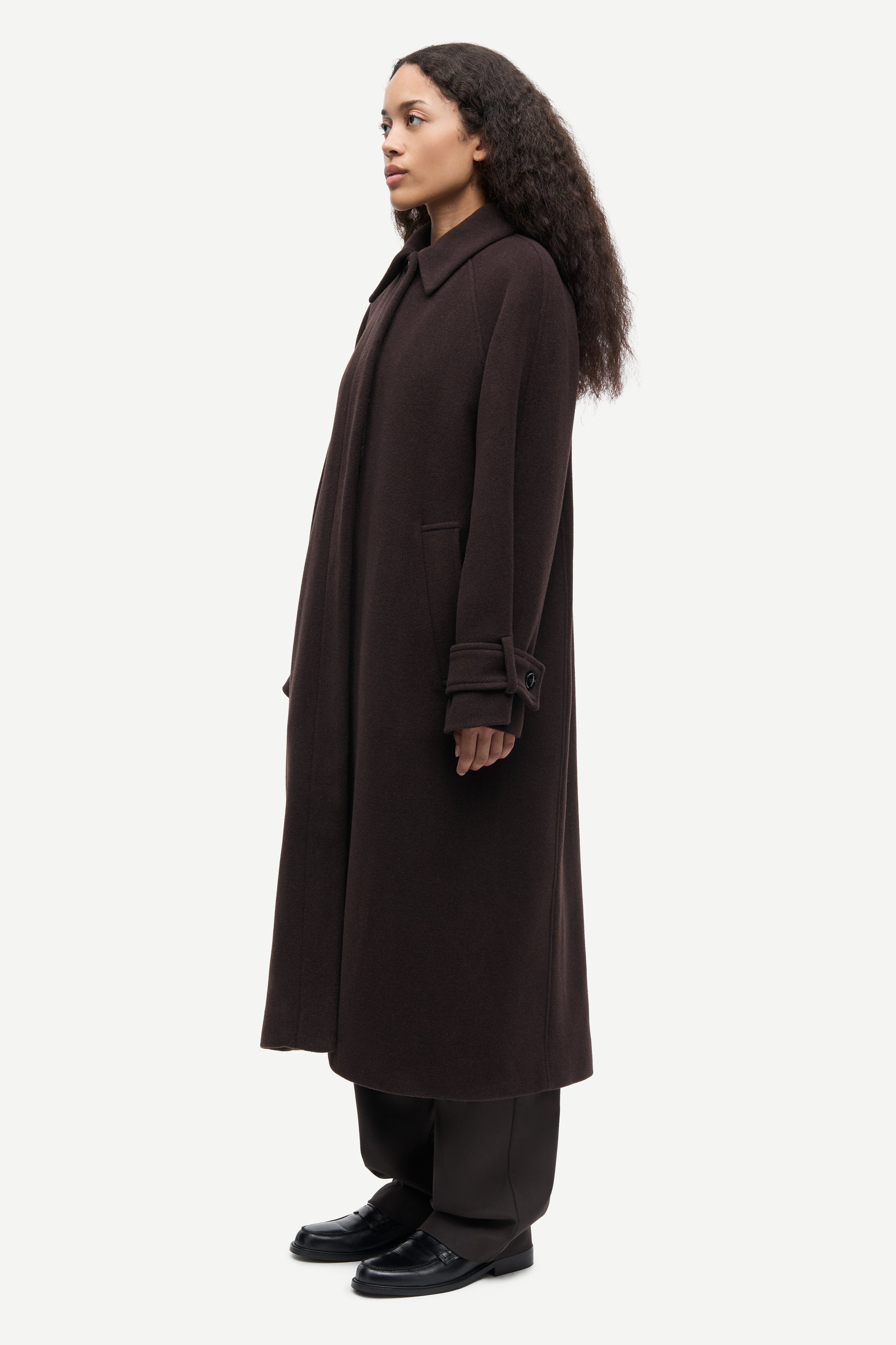 A woman modeling a long, dark brown wool-blend coat with matching dark trousers and black shoes, shown in profile against a light gray studio background.