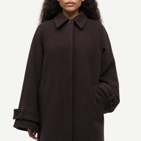 A woman wearing a dark brown wool-blend coat with a collar, front button closure, and large patch pockets, photographed against a light gray studio background.