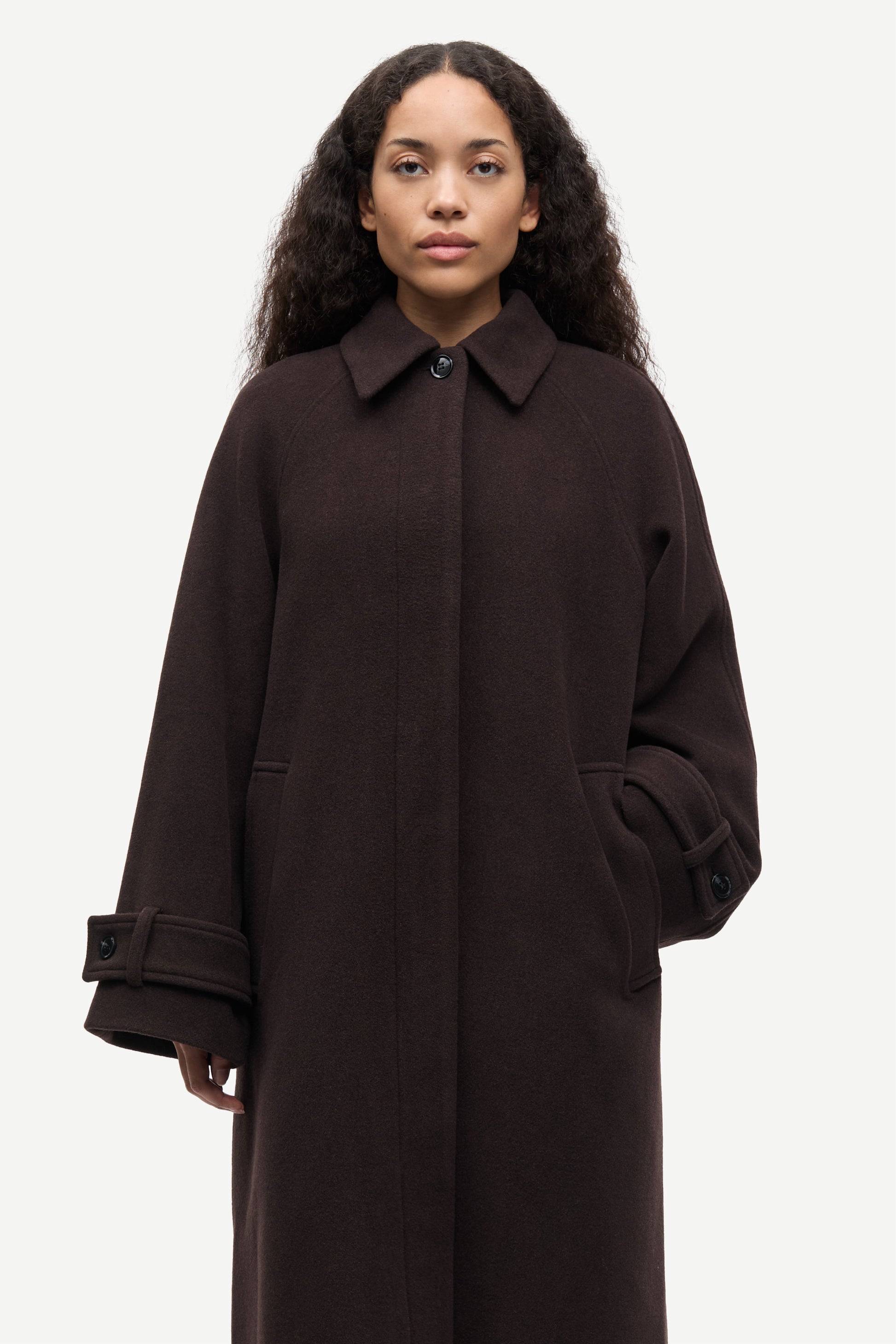 A woman wearing a dark brown wool-blend coat with a collar, front button closure, and large patch pockets, photographed against a light gray studio background.