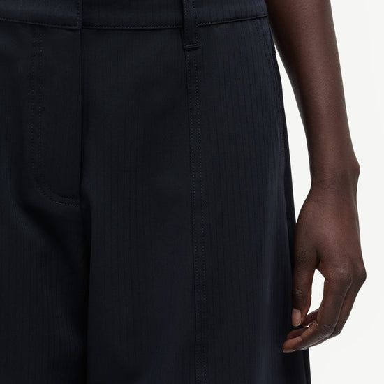 Close-up of a person wearing dark navy pinstriped trousers, showing the waistband, front pocket, and hand against a light neutral background.