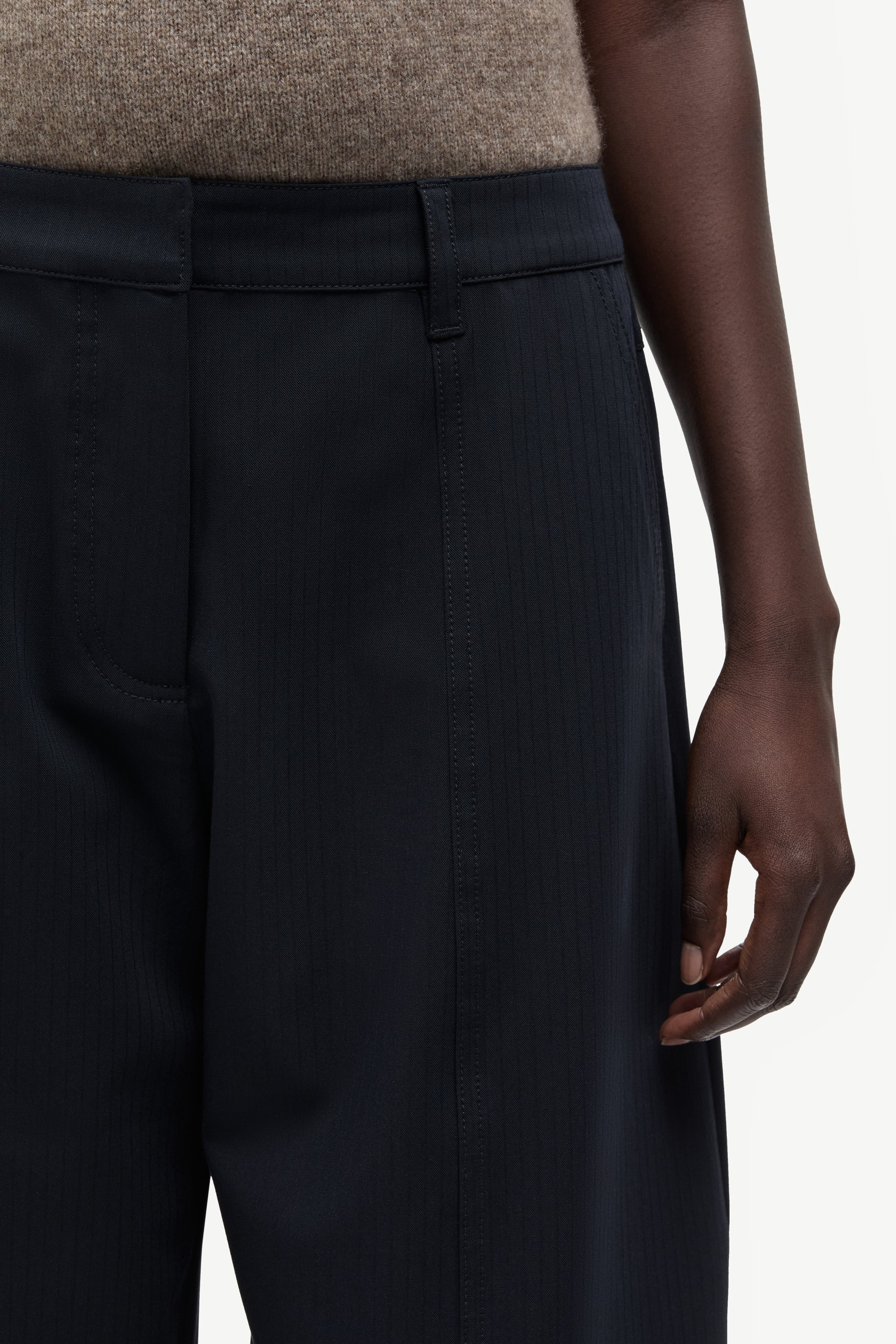 Close-up of a person wearing dark navy pinstriped trousers, showing the waistband, front pocket, and hand against a light neutral background.