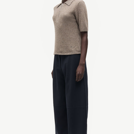A model stands in profile wearing a short-sleeve knit polo shirt in a light taupe-beige color and wide-leg dark navy trousers, photographed against a clean off-white studio background.