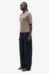 A model stands in profile wearing a short-sleeve knit polo shirt in a light taupe-beige color and wide-leg dark navy trousers, photographed against a clean off-white studio background.
