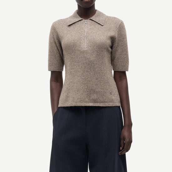 A model wearing a beige short-sleeve knit polo shirt with a collar and dark navy wide-leg trousers, photographed against a solid light gray background.