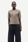 A model wearing a beige short-sleeve knit polo shirt with a collar and dark navy wide-leg trousers, photographed against a solid light gray background.