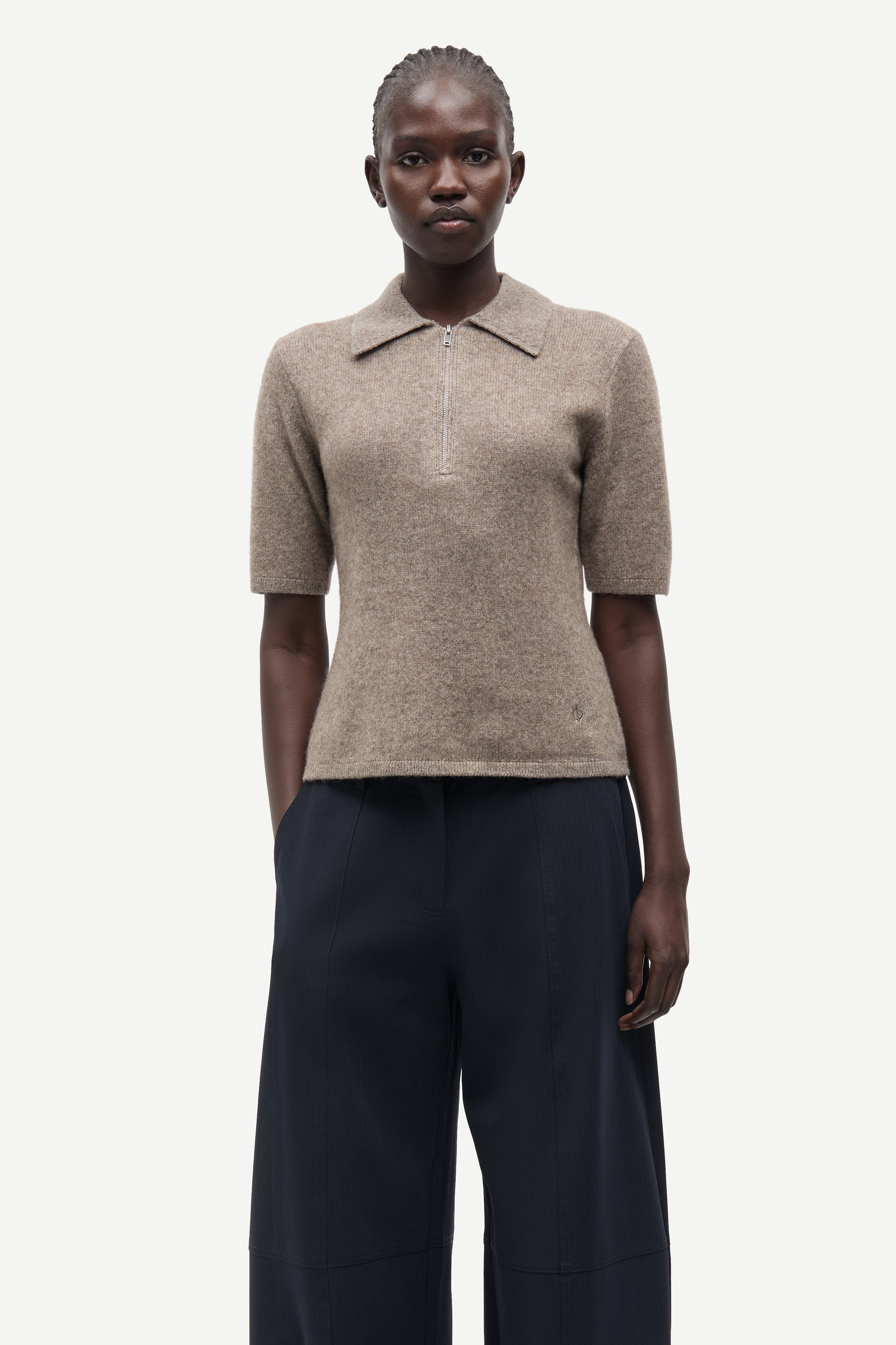A model wearing a beige short-sleeve knit polo shirt with a collar and dark navy wide-leg trousers, photographed against a solid light gray background.