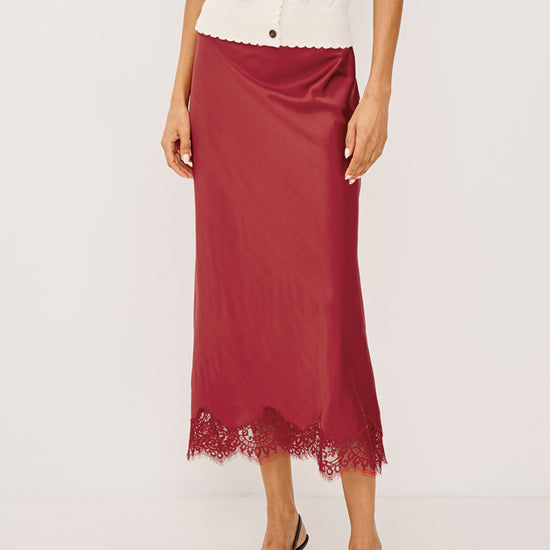 A model is shown from the chest down wearing a dusty rose satin midi skirt with a lace hem, paired with a white argyle cardigan and black pointed-toe heels.
