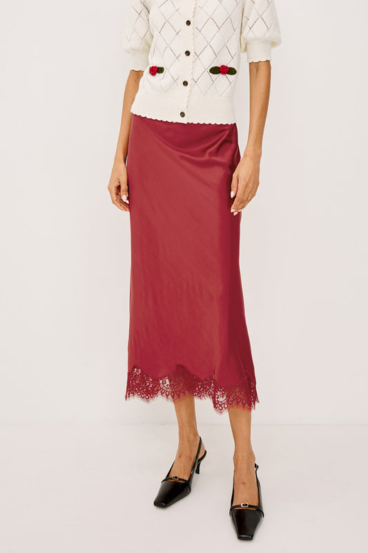 A model is shown from the chest down wearing a dusty rose satin midi skirt with a lace hem, paired with a white argyle cardigan and black pointed-toe heels.