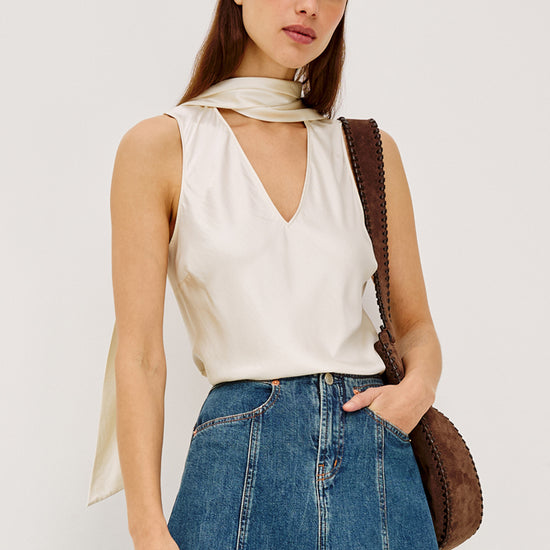 Woman wearing a white sleeveless top and blue jeans with a brown leather bag on a plain background