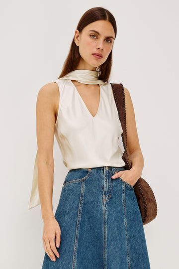 Woman wearing a white sleeveless top and blue jeans with a brown leather bag on a plain background