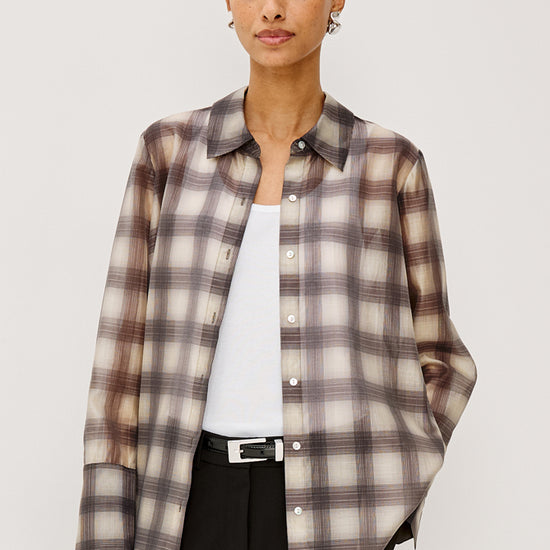 A woman modeling a long-sleeve button-up plaid shirt in beige, brown, and gray tones, worn open over a white top and black trousers, against a light gray studio background.