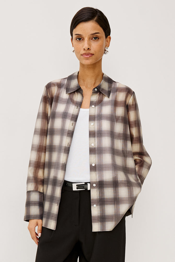 A woman modeling a long-sleeve button-up plaid shirt in beige, brown, and gray tones, worn open over a white top and black trousers, against a light gray studio background.