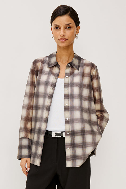 A woman modeling a long-sleeve button-up plaid shirt in beige, brown, and gray tones, worn open over a white top and black trousers, against a light gray studio background.