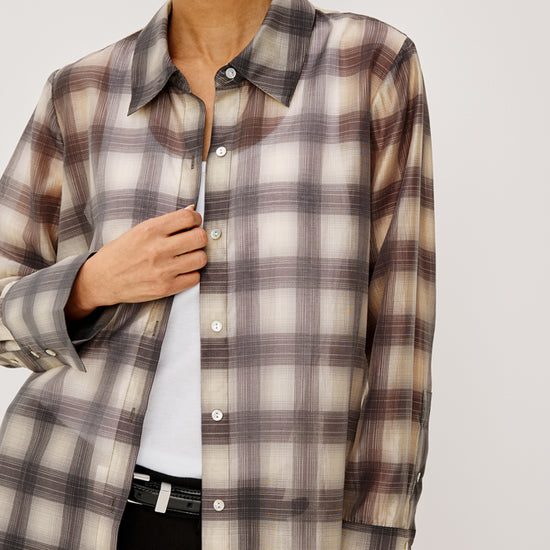 A woman wearing a long-sleeve button-up shirt with a brown, beige, and gray plaid pattern over a white top and black pants, shown against a light gray studio background.