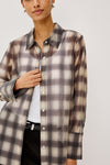 A woman wearing a long-sleeve button-up shirt with a brown, beige, and gray plaid pattern over a white top and black pants, shown against a light gray studio background.