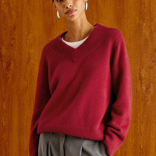 A woman wearing a burgundy V-neck knit sweater over a white undershirt and gray trousers, standing against a warm wood-paneled background.