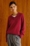 A woman wearing a burgundy V-neck knit sweater over a white undershirt and gray trousers, standing against a warm wood-paneled background.